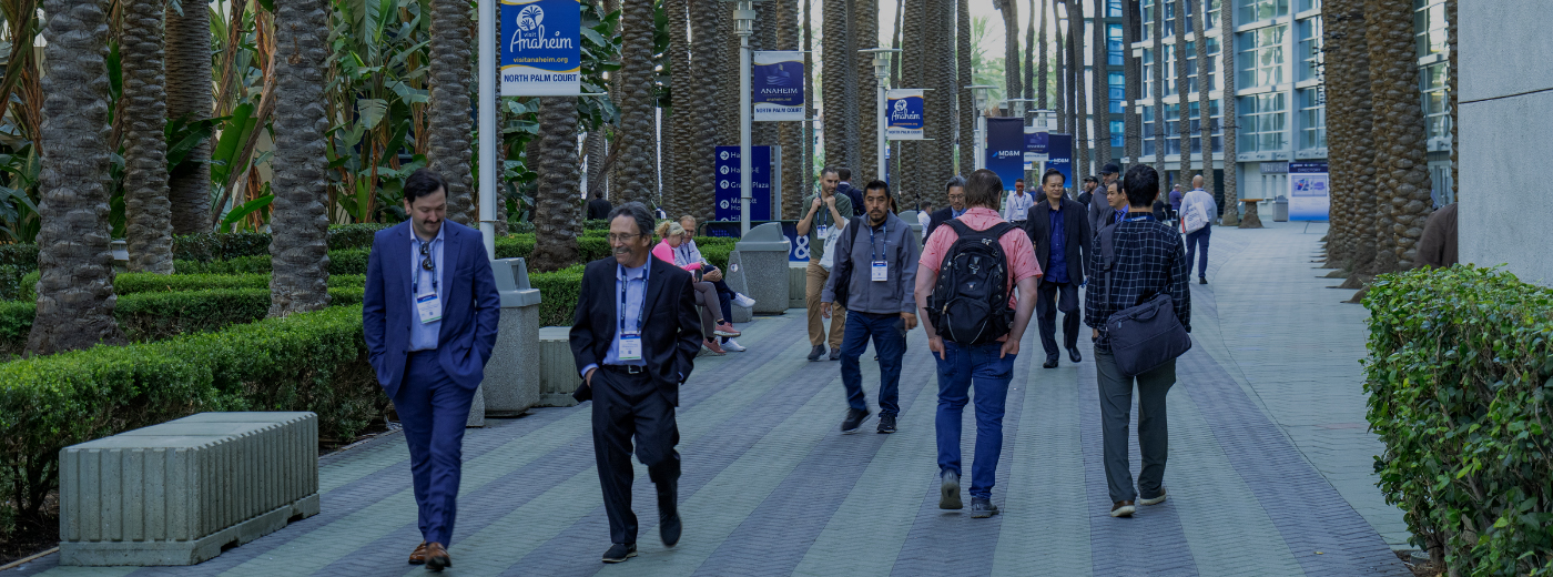 A diverse group of people walks down a palm tree-lined sidewalk outside of Anaheim Convention Center