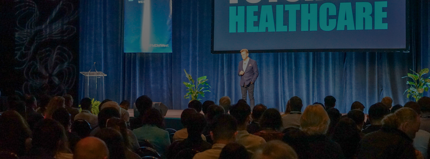 A speaker stands on stage at a conference, addressing the audience with a large screen displaying visuals behind him