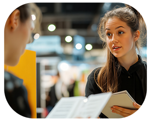 At a trade show, one woman speaks to another, with a vibrant atmosphere filled with displays and visitors around them