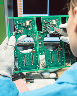 A woman in a blue shirt holds a green circuit board, examining its components with a focused expression