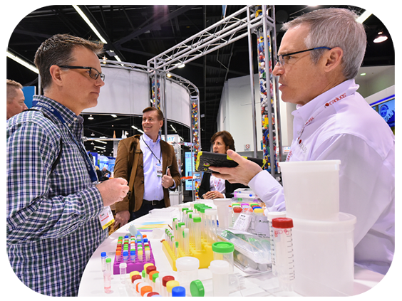 Two men converse at a trade show, with numerous vendor booths and attendees visible in the background